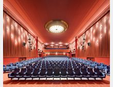 Ein Kino- oder Theatersaal mit roten Wänden und einem großen, runden Deckenlicht in der Mitte. Die Sitzreihen bestehen aus blauen Polstersitzen mit goldenen Details, und an den Wänden sind Beleuchtungen sowie Lautsprecher angebracht.