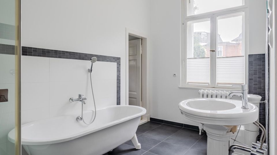 Helles Badezimmer mit freistehender Badewanne an einer weiß gefliesten Wand mit dunkler Mosaikbordüre, großem Fenster und dunkelgrauen Bodenfliesen. Im Vordergrund stehen ein rundes Waschbecken auf Säule und ein Heizkörper unter dem Fenster.