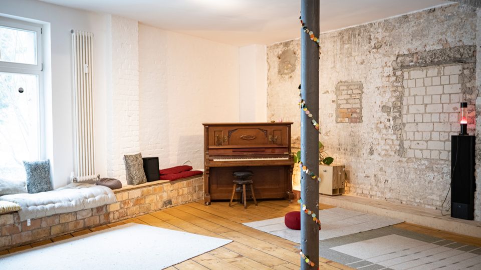 Heller Innenraum im Loft-Stil mit Holzfußboden, weiß gestrichenen Ziegelwänden und einer freigelegten, teils verputzten Backsteinwand. An der Fensterseite steht eine gepolsterte Sitzbank mit Kissen; in der Mitte ein Klavier mit Hocker sowie ein Metallstützpfeiler, daneben Teppiche und ein Lautsprecher.