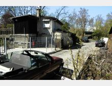 Eine ländliche Straße mit Kopfsteinpflaster, gesäumt von einfachen Holzhäusern und einem Maschendrahtzaun. Im Vordergrund steht ein rotes Cabriolet, während im Hintergrund mehrere Autos und Bäume zu sehen sind.