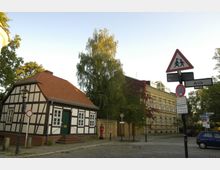 Fachwerkhaus mit rotem Ziegeldach neben einem historischen, mit Efeu bewachsenen Gebäude; im Vordergrund eine Kopfsteinpflasterstraße mit einem Straßenschild und mehreren Verkehrsschildern. Im Hintergrund Bäume unter klarem Himmel.