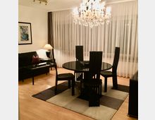 Modernes Wohn- und Esszimmer mit rundem schwarzem Esstisch und vier hohen Stühlen auf einem karierten Teppich, darüber ein großer Kristalllüster. Im Hintergrund bodentiefe Fenster mit weißen Vorhängen, links ein schwarzes Sofa mit Couchtisch, Stehlampe und gerahmtem Wandbild.