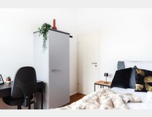 Helles Schlafzimmer mit einem weißen Kleiderschrank, einem schwarzen Schreibtisch mit Stuhl und einer Dekopflanze. Rechts befinden sich ein Bett mit Kissen in Schwarz und Gold sowie ein kleiner Beistelltisch mit einer Lampe.