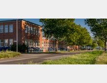Backstein-Gebäude mit großen, weißen Fenstern und einem Schild mit der Aufschrift "Jobcenter" an der Fassade. Vor dem Gebäude verläuft eine von Bäumen gesäumte Straße, umgeben von Grünflächen.
