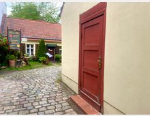 Ein kleiner Innenhof mit einem Kopfsteinpflasterweg, flankiert von einem Gebäude mit beiger Fassade und einer roten Tür im Vordergrund. Im Hintergrund ein niedrigeres Gebäude mit roten Dachziegeln, weißen Fenstern und einem Schild mit der Aufschrift „GEWERBE HOF“, umgeben von Pflanzen und Dekorationen.