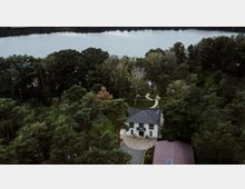 Luftaufnahme eines weißen zweistöckigen Hauses mit dunklem Schrägdach, umgeben von dichtem Wald. Im Hintergrund ist ein großer See sichtbar, der von Bäumen eingerahmt ist, sowie gekrümmte Gehwege und eine Grünfläche.