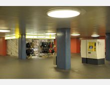 Eine unterirdische Passage mit einem Kiosk für Reisebedarf, der Taschen und andere Waren anbietet. Der Raum ist durch blaue, mit Fliesen verkleidete Säulen, rote Wände und helle Deckenleuchten geprägt; daneben steht ein gelber Fahrkartenautomat.
