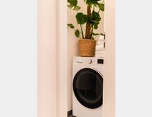 Eine moderne weiße Waschmaschine in einer schmalen Nische, daneben gestapelte weiße Handtücher und eine große grüne Pflanze in einem geflochtenen Korb.