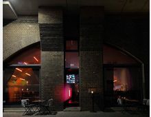 Backsteinfassade eines Gebäudes mit großen, bogenförmigen Fenstern, die in einen beleuchteten Innenraum mit modernen Neonlichtern und dekorativen Möbeln blicken. Davor stehen einige kleine Tische und Stühle im Freien auf einem gepflasterten Gehweg, teils mit Decken über den Stühlen.