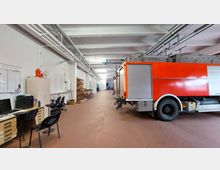 Eine geräumige Feuerwehrgarage mit einem roten Feuerwehrfahrzeug auf der rechten Seite. Links stehen Büromöbel mit Schreibtischen, Stühlen und Regalen, während die Decke mit Lüftungs- und Rohrleitungen versehen ist.