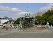 Brunnen mit einer zentralen Figur des Neptun und mehreren umliegenden Meerjungfrauen aus Bronze; Wasser strömt aus verschiedenen Elementen der Skulptur. Im Hintergrund befinden sich Bäume, ein modernes Gebäude mit auffälliger Architektur und ein mehrstöckiges hohes Wohn- oder Bürogebäude.