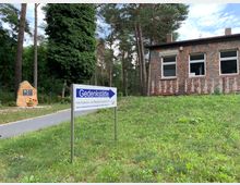 Ein Gedenkort mit einem Wegweiser „Gedenkstätte“, einem gepflasterten Weg, einem Gedenkstein mit Blumen und einem einfachen Backsteingebäude im Hintergrund, umgeben von Bäumen und Grünflächen.