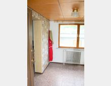 Ein kleiner Raum mit einer Wand aus gemusterter Tapete, einem Fenster mit Holzrahmen, einem Heizkörper darunter und einer Decke mit Holzpaneelen. An der Wand hängt ein roter und beiger Mantel. Der Boden ist mit rötlichen Fliesen ausgelegt.