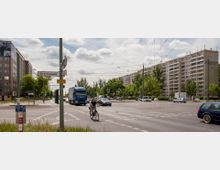Straßenkreuzung in einer Stadt mit einem Fahrradfahrer im Vordergrund, umgeben von mehreren Fahrspuren, Autos, einem Lkw und einem Plattenbau im Hintergrund. Straßenlaternen, Oberleitungen für Straßenbahnen und Straßenbeschilderung sind sichtbar, darunter ein Schild mit der Aufschrift „Allee der Kosmonauten“.