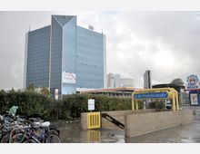 Ein modernes Hochhaus mit blauer Glasfassade steht neben einer Tankstelle mit einem „Sprint“-Logo. Im Vordergrund ist ein Eingang zur U-Bahn-Station „Paulsternstraße“, umgeben von Fahrrädern und nassem Gehweg.