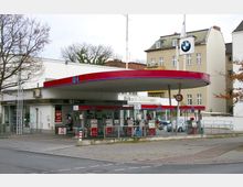 Tankstelle mit rotem Dach und mehreren Zapfsäulen, darauf die Aufschrift „Q1“. Im Hintergrund sind ein kleines Geschäft, der Schriftzug „Riller & Schnauck“ sowie mehrstöckige Wohngebäude zu sehen.