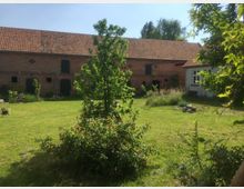 Ein weitläufiger grasbewachsener Garten mit einem kleinen Baum in der Mitte. Im Hintergrund ist ein großes, altes Backsteingebäude mit roten Ziegeldächern und grünen Türen zu sehen, flankiert von einem kleineren Wohngebäude rechts.