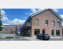 Zweistöckiges Gebäude aus roten Backsteinen mit Geschäften im Erdgeschoss, darunter eine Bäckerei. Im Vordergrund ein Parkplatz mit einem Behindertenparkplatzschild und ein geparkter schwarzer Wagen, daneben ein Gehsteig und einige Sitzgelegenheiten.