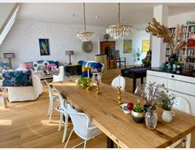 Offener Wohnraum mit modernem und gemütlichem Interieur. Ein großer Holztisch steht im Vordergrund, geschmückt mit Vasen und Kerzen, während im Hintergrund bunte Sofas, ein Bücherregal und Dekorelemente wie Kronleuchter und Kunst an weißen Ziegelwänden zu erkennen sind.