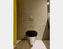 Minimalistischer Innenraum mit glatter grauer Wand, mittig montiertem Druckknopf und einem kleinen zylindrischen Metallhocker mit schwarzer Sitzfläche auf einem hell gesprenkelten Boden. Links ist ein gelber Vorhang zu sehen, rechts eine spiegelnde Fläche mit Leuchte.