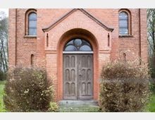 Fassade eines Gebäudes aus rotem Backstein mit einem hölzernen Doppeltor. Über der Tür befindet sich ein halbrundes Fenster, umrahmt von einem Ziegelbogen, und die Umgebung ist von Sträuchern und Bäumen geprägt.