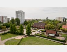 Luftaufnahme einer Stadtlandschaft mit einem Mix aus hohen Wohngebäuden und einem kleineren Bauernhofareal im Vordergrund. Der Bauernhof ist umgeben von grünen Wiesen, Bäumen und Wegen, während im Hintergrund moderne Wohnhochhäuser in einer grünen Umgebung stehen.
