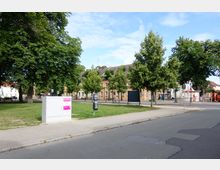 Straßenansicht mit Gehweg, Rasenfläche und Bäumen im Vordergrund. Im Hintergrund stehen beige Wohngebäude mit roten Ziegeldächern, daneben sind Straßenschilder, Fahrräder und ein orangefarbener Lastwagen zu sehen.