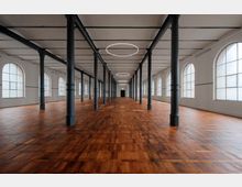 Großer, leerer Raum mit dunklem Holzboden, Reihen von schwarzen Metallstützpfeilern und einem weißen Deckensystem. Die Wände haben große, bogenförmige Fenster, die viel Licht einfallen lassen, und runde, minimalistische Deckenleuchten ziehen sich durch den gesamten Raum.