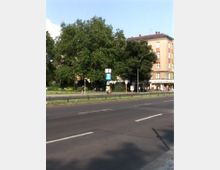 Straßenansicht mit einer mehrspurigen Fahrbahn, angrenzendem Grünstreifen und großen Bäumen im Hintergrund. Rechts ist ein mehrstöckiges Wohngebäude mit weißer und roter Fassade sowie einem Geschäft im Erdgeschoss sichtbar.