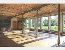 Großer, heller Raum mit einer Decke aus freiliegenden Ziegeln, Metallstützpfeilern und großen Glasfenstern, die auf eine grüne Gartenlandschaft blicken. Auf der linken Seite befinden sich eine offene Küche, ein Esstisch mit Stühlen und Holzschränke.