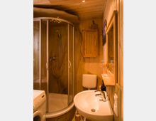 Kleines Badezimmer mit Holzwänden und -decke sowie einer runden Duschkabine mit Glastüren. Ausstattung umfasst ein Waschbecken, eine Toilette, einen Wandschrank aus Holz, einen Spiegel und eine Waschmaschine.