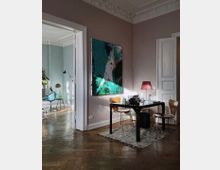 Helles Wohnzimmer mit Parkettboden und Stuckverzierungen an der Decke, ausgestattet mit einem schwarzen Tisch und zwei Holzstühlen. An der Wand hängt ein großes, abstraktes Gemälde eines Vogels, daneben steht eine Tischlampe mit rotem Lampenschirm.