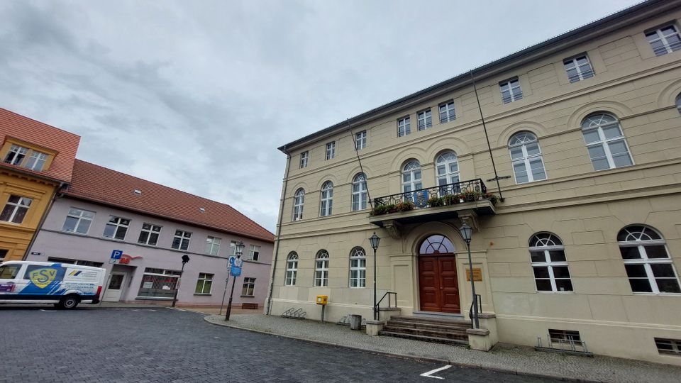 Historisches Gebäude mit hellbeiger Fassade, symmetrischen Rundbogenfenstern und einem Eingang mit einem roten Holztor, darüber ein Balkon mit Blumenschmuck. Rechts und links stehen Straßenlaternen, im Hintergrund befinden sich weitere Gebäude mit roten Ziegeldächern.