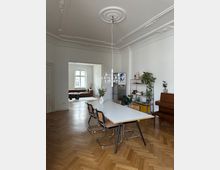 Esszimmer in einem Altbau mit Fischgrätenparkett, einem weißen Tisch mit vier geflochtenen Stühlen und einer hängenden Deckenlampe. Im Hintergrund ist ein Wohnzimmer mit einem braunen Sofa sichtbar, Deckenstuck und helle Wände prägen das Ambiente.