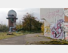 Ein verlassenes Gelände mit einem zylindrischen Backsteinturm, der eine Kuppel und eine umlaufende Plattform trägt, sowie einem angrenzenden Gebäude mit Wellblechwänden, die mit Graffiti bedeckt sind. Der Boden ist teils asphaltiert, teils überwuchert, und im Hintergrund sind Bäume und verwilderte Vegetation erkennbar.