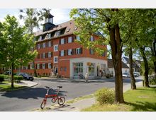 Mehrstöckiges Wohngebäude mit orangefarbener Fassade und weißen Fensterrahmen, das an einer Straßenecke steht. Im Erdgeschoss befinden sich ein kleiner Laden mit Lotto-Logo und Eingänge, umgeben von Bäumen, Gehwegen und parkenden Autos.