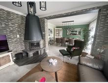 Wohnzimmer mit gemauertem Kamin in dunkler Optik und dekorativer Steinwand, verbunden mit einem Essbereich. Im Raum befinden sich moderne Möbel, darunter ein grüner Sessel, ein Ledersofa, ein Esstisch mit Stühlen und eine große Wandspiegel-Dekoration.