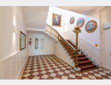 Historisches Treppenhaus mit gemustertem Boden aus braunen und weißen Fliesen, einer weißen Holzvertäfelung und einer Treppe mit rotem Teppich und goldgeländer. An den Wänden befinden sich Gemälde und dekorative runde Rahmen.