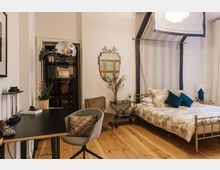 Schlafzimmer mit Holzboden, einem Metallbett mit gemusterten Kissen und Bettwäsche, einem Schreibtisch mit Lampe und Stuhl sowie dekorativen Elementen wie einem Spiegel an der Wand, Pflanzen und Beistelltischen. Ein Baldachin mit gestreiftem Stoff hängt über dem Bett, während Tageslicht durch ein Fenster mit Vorhängen einfällt.