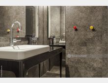 Modernes Badezimmer mit Waschbecken auf dunklem Waschtisch, glänzender Armatur und hellen Wänden in grauer Marmoroptik. Im Hintergrund befinden sich Haken in Rot und Gelb sowie ein großer Spiegel.