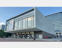 Modernes Kino mit der Aufschrift „International“ auf der Fassade, einem großen Banner zu einem Festival sowie Glasfronten über dem Eingangsbereich. Die seitliche Wand zeigt ein kunstvolles Relief, flankiert von einem mehrstöckigen Gebäude im Hintergrund.