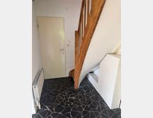 Kleiner Innenflur mit einer geschlossenen, hellen Tür und einer Holztreppe mit Geländer; links steht ein Heizkörper an der Wand. Der Boden ist mit unregelmäßigem, dunklem Steinmosaik gefliest.