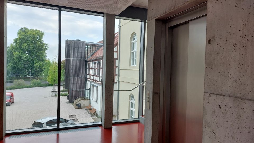 Blick durch eine große Glasfront auf einen Innenhof mit Pflastersteinen, umgeben von einem historischen Fachwerkgebäude und einem modernen Anbau. Rechts im Vordergrund befindet sich eine Metall-Aufzugstür in einer Betonwand.