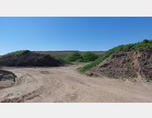 Offene Fläche mit sandigem Boden, auf dem Spuren von Fahrzeugen zu erkennen sind. Umgeben von Erdhügeln und bewachsenen grasigen Erhebungen, mit einem klaren blauen Himmel im Hintergrund.