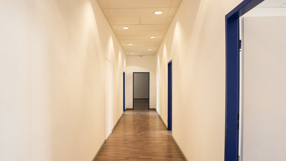 Langer, schmaler Innenflur in einem Büro- oder Verwaltungsgebäude mit weißen Wänden, Holzlaminatboden und abgehängter Decke mit runden Einbauleuchten. Entlang des Gangs befinden sich mehrere Türen mit blauen Rahmen, am Ende ist ein offener Durchgang zu einem weiteren Raum sichtbar.