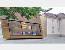 Moderner Anbau mit goldenen Fassadenplatten und einer großen Glasfront, durch die eine bunte Inneneinrichtung mit Bücherregalen sichtbar ist, daneben ein historisches Gebäude mit hell verputzter Fassade und roten Dachziegeln. Ein Baum mit grünem Laub ragt in das Bild hinein.