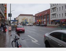 Straßenszene an der Hermannstraße in Berlin mit mehrstöckigen Wohnhäusern und einer Reihe von Geschäften, darunter ein Rewe- und ein NKD-Markt in einem roten Backsteingebäude. Im Vordergrund stehen geparkte Autos und Fahrräder, Fußgänger laufen auf dem Gehweg, und eine U-Bahn-Station ist im Hintergrund erkennbar.