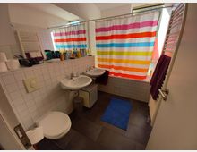 Ein Badezimmer mit einer Duschwanne, die durch einen Duschvorhang mit bunten Streifen abgetrennt ist. Es gibt zwei Waschbecken mit Spiegel darüber, daneben eine weiße Toilette und ein Handtuchhalter mit lilafarbenen Handtüchern; der Boden ist mit dunklen Fliesen ausgelegt.