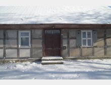 Fassade eines niedrigen Fachwerkhauses mit verschneitem Dach, mittiger dunkler Holztür mit zwei Stufen und zwei Fenstern mit Holzläden. Davor liegt eine schneebedeckte Fläche am Gebäude.