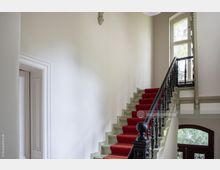 Treppenhaus in einem historischen Gebäude mit einer roten Teppichtreppe, weißen Wänden und verzierten schwarzen Holzgeländern. Rechts oben befindet sich ein großes Fenster, das natürliches Licht hereinlässt.
